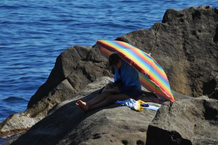 Алупка.  Женщина сидит на камнях на берегу моря под зонтом.