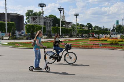 Москва.  Девушки на велосипеде и самокате на дорожках ВДНХ.