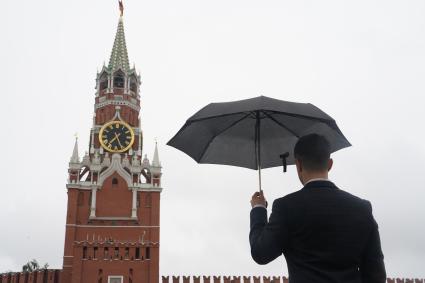 Москва.  Мужчина в костюме  под зонтом на Красной площади смотрит на Спасскую башню Кремля.