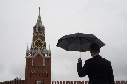 Москва.  Мужчина в костюме  под зонтом на Красной площади смотрит на Спасскую башню Кремля.