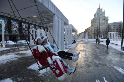 Москва.   Мужчина и женщина в костюмах деда Мороза и Снегурочки на качелях на Триумфальной площади.