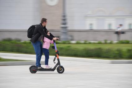 Москва.  Мужчина с девочкой едет на электросамокате  на ВДНХ.