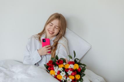 Красноярск. Девушка с мобильным телефоном и букетом цветов лежит в постели.