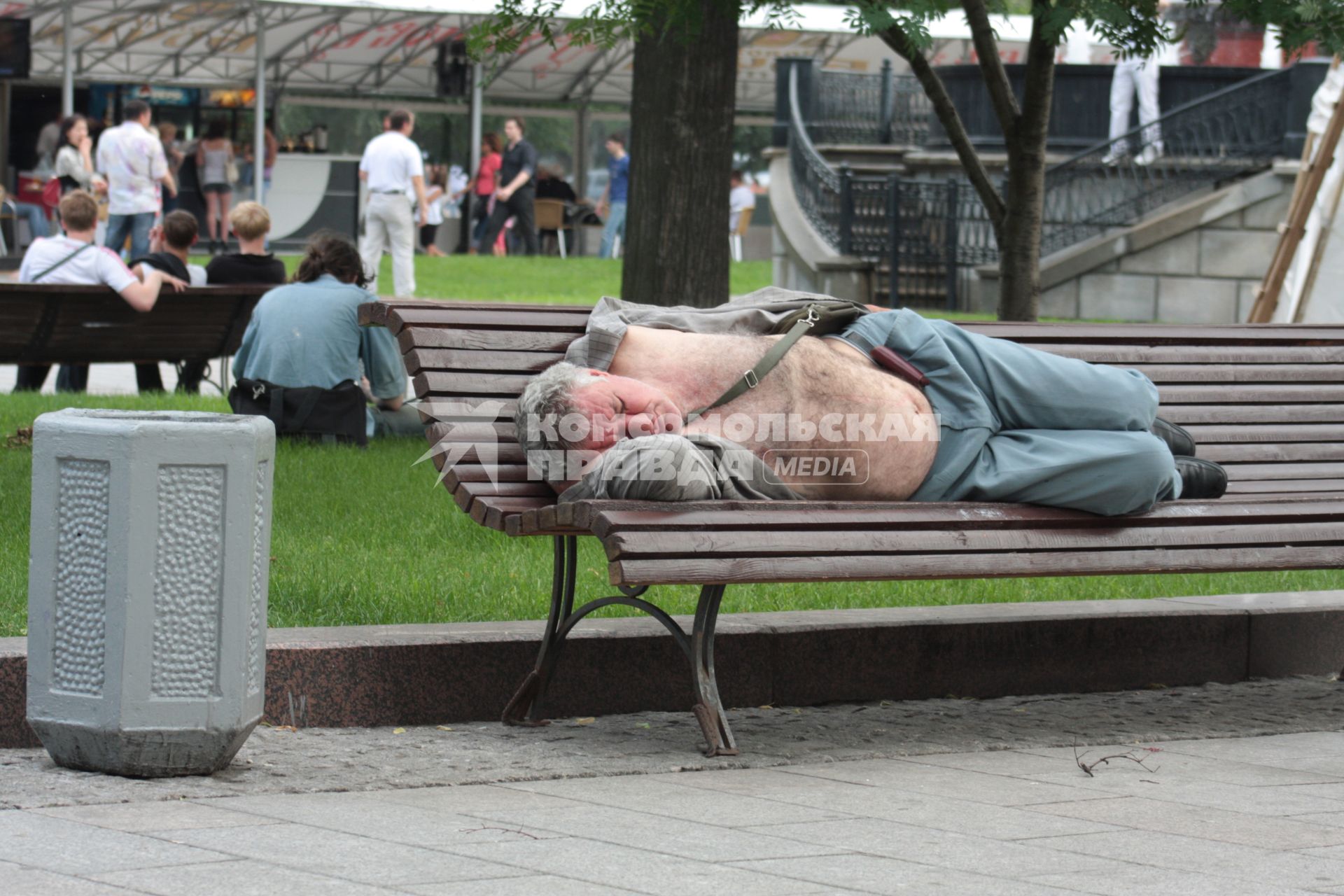 Спящий бомж днем лежит на скамейке в сквере.