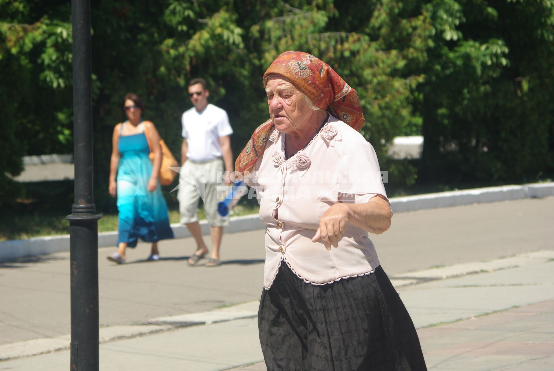 Пожилая женщина с палочкой идёт по городу, на заднем фоне молодая пара мужчина и женщина, 21 июля 2010 года. Старость и молодость, пенсия, пенсионная реформа, зарплата, старушка, бабка, старуха, молодые, будущее, семья, дорога жизни, город, лето, косынка,