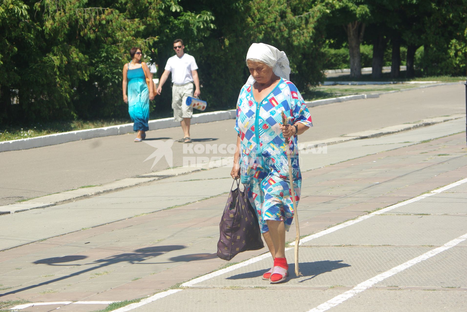 Пожилая женщина с палочкой идёт по городу, на заднем фоне молодая пара мужчина и женщина, 21 июля 2010 года. Старость и молодость, пенсия, пенсионная реформа, зарплата, старушка, бабка, старуха, молодые, будущее, семья, дорога жизни, город, лето, косынка,