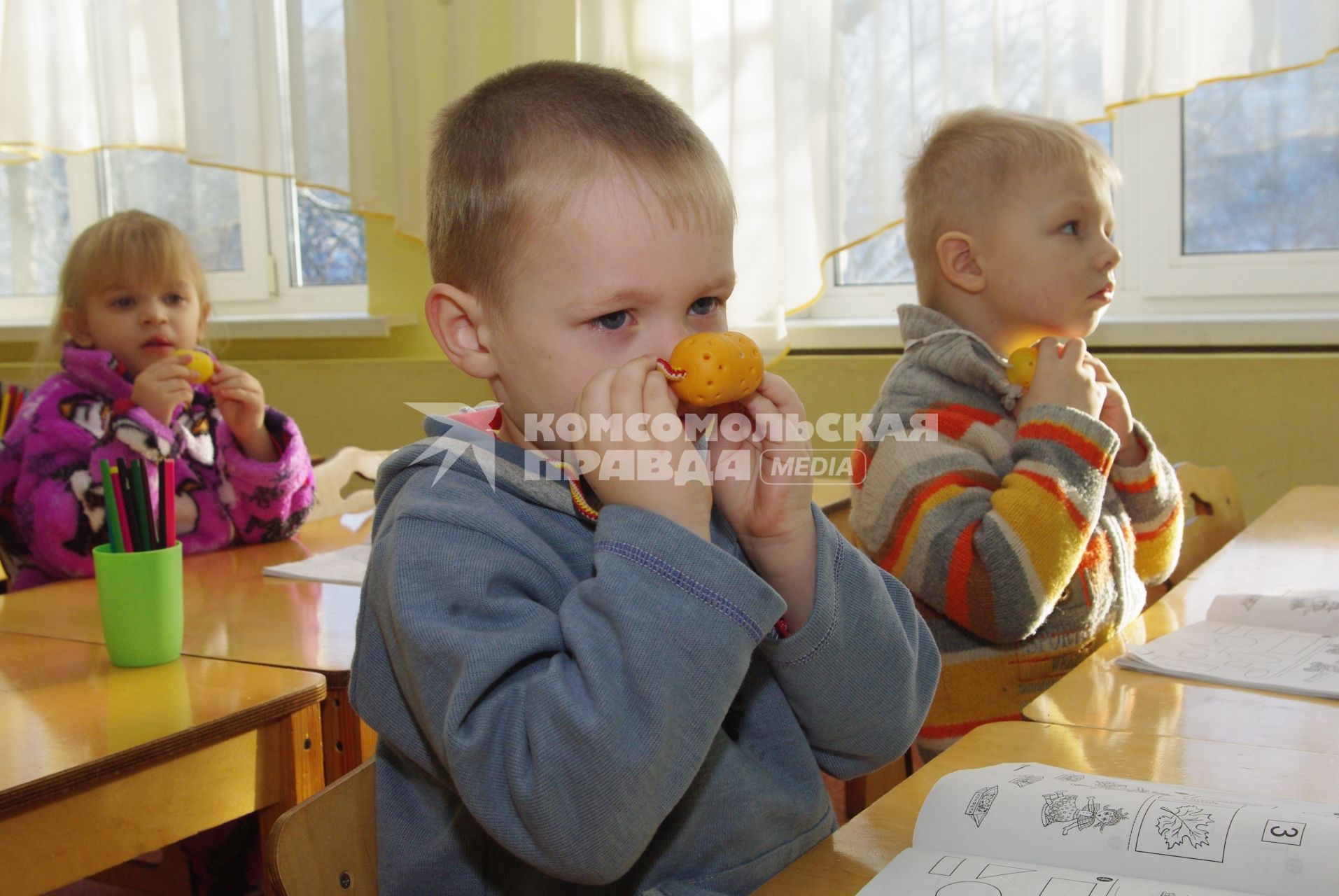 Дети в детском саду сидят за партами и нюхают чеснок и лук, которые лежат в киндер- сюрпризах с дырочками и висят на шеях детей., Саратов, 3 февраля 2011 года. Детсад, детский сад, дети, воспитатель, занятия, обучение, парта, профилактика гриппа, грипп, болезнь, каратнин.