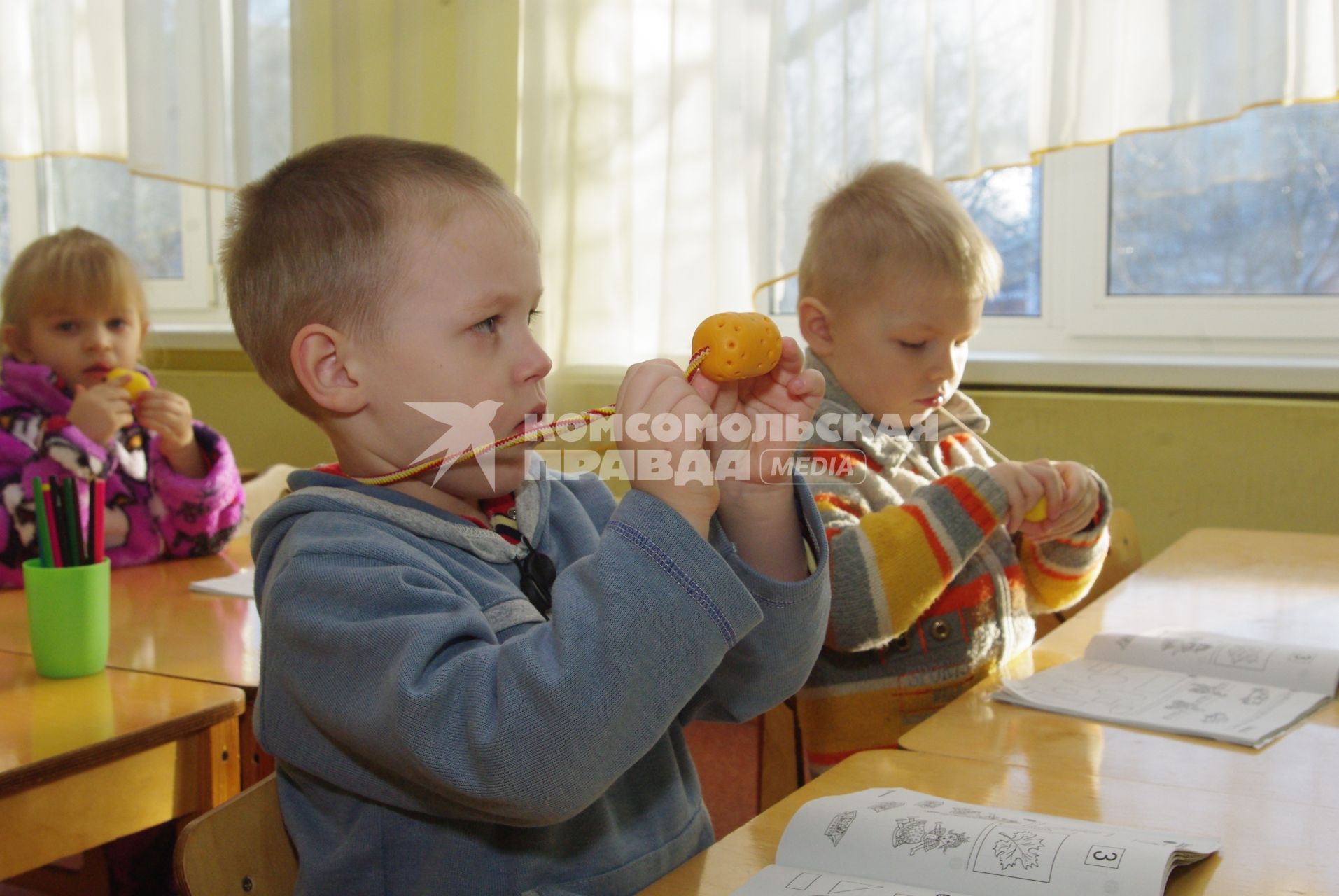 Дети в детском саду сидят за партами и нюхают чеснок и лук, которые лежат в киндер- сюрпризах с дырочками и висят на шеях детей., Саратов, 3 февраля 2011 года. Детсад, детский сад, дети, воспитатель, занятия, обучение, парта, профилактика гриппа, грипп, болезнь, каратнин.