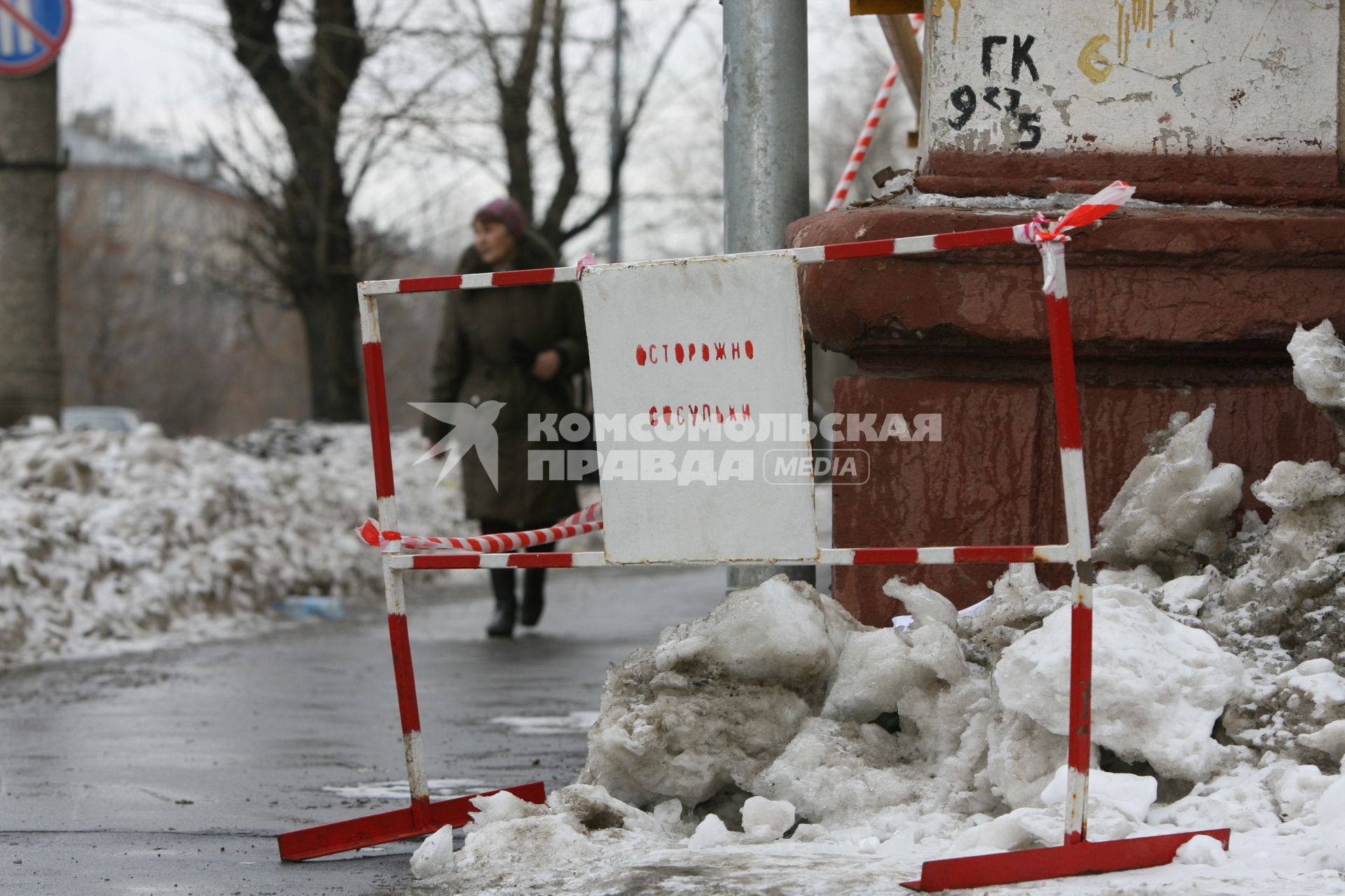 На углу дома стоит табличка с надписью- ОСТОРОЖНО СОСУЛЬКИ мимо проходит женщина. 3 марта 2011 года.