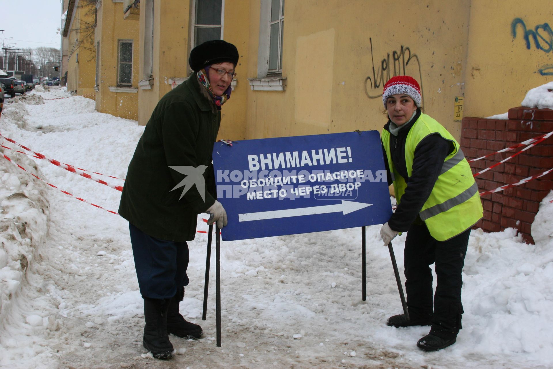 Две женщины перетаскивают указатель с надписью ВНИМАНИЕ! ОБОЙДИТЕ ОПАСНОЕ МЕСТО ЧЕРЕЗ ДВОР. Уфа. 17 марта 2011 года.