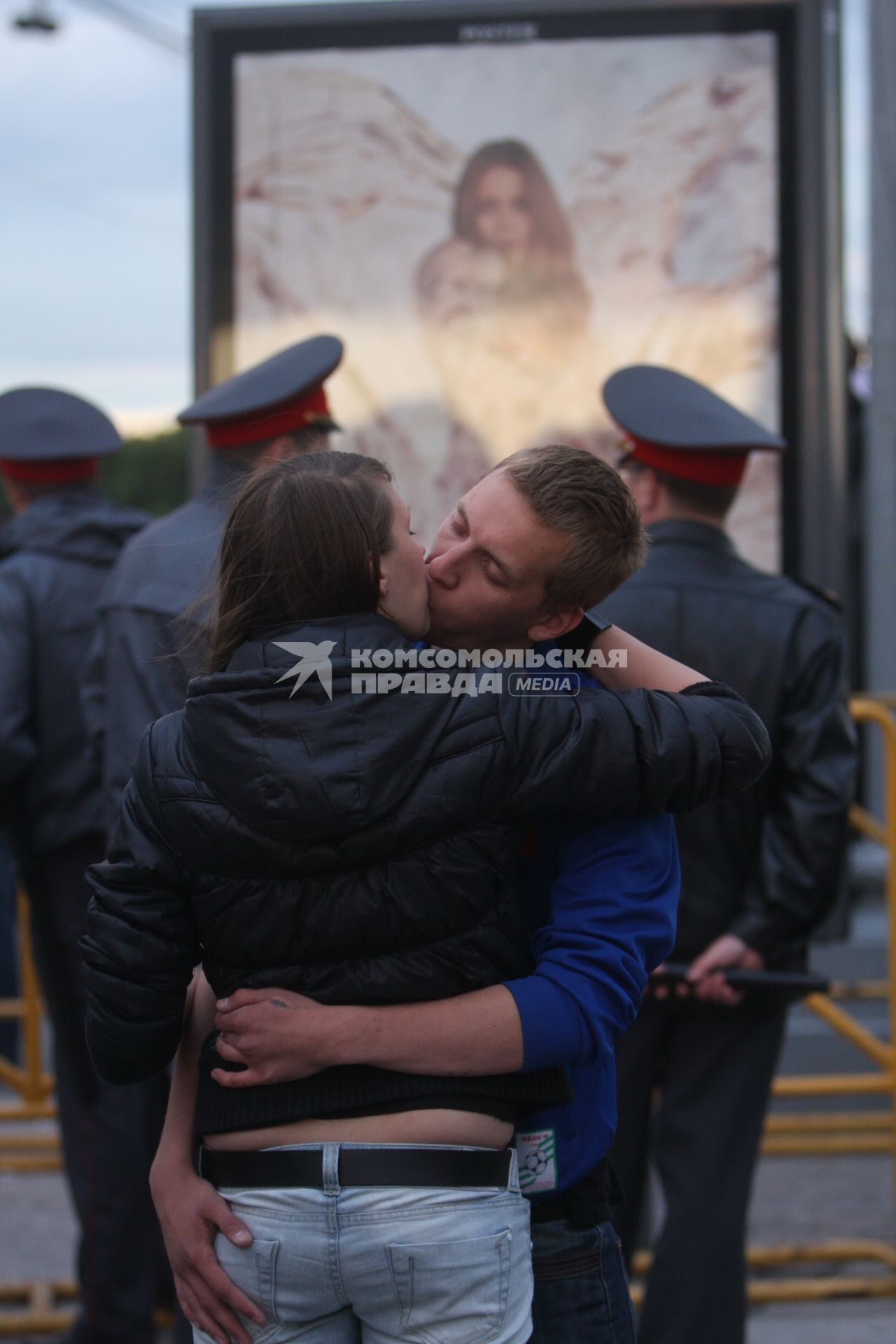 Мужчина и женщина целуются на улице. На заднем плане стоят представители охраны правопорядка (полицейские).