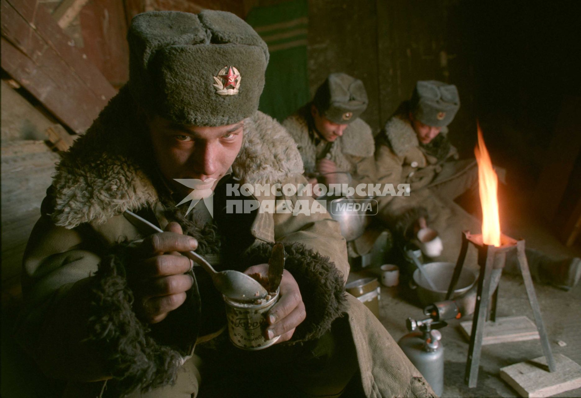 На переднем плане солдат в зимнем полушубке ест ложкой тушенку прямо из банки.На заднем плане солдаты сидя на полу готовят пищу на керосиновой горелке.