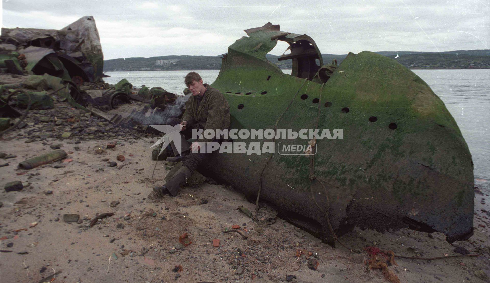 Человек в камуфлированном  бушлате сидит на ржавых остатках военного корабля на берегу моря.