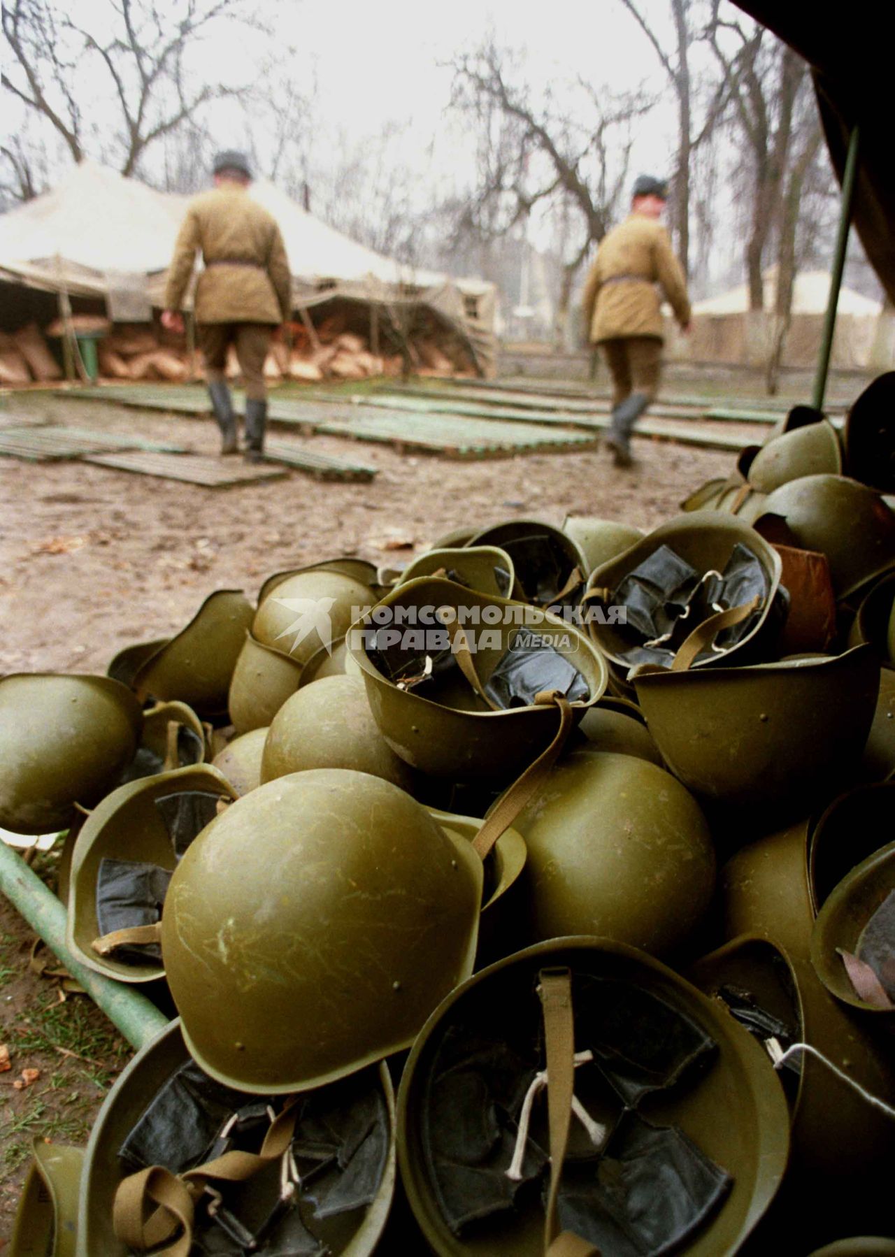 На переднем плане сваленные в кучу военные каски.На заднем плане двое солдат идут к палаткам военного образца.