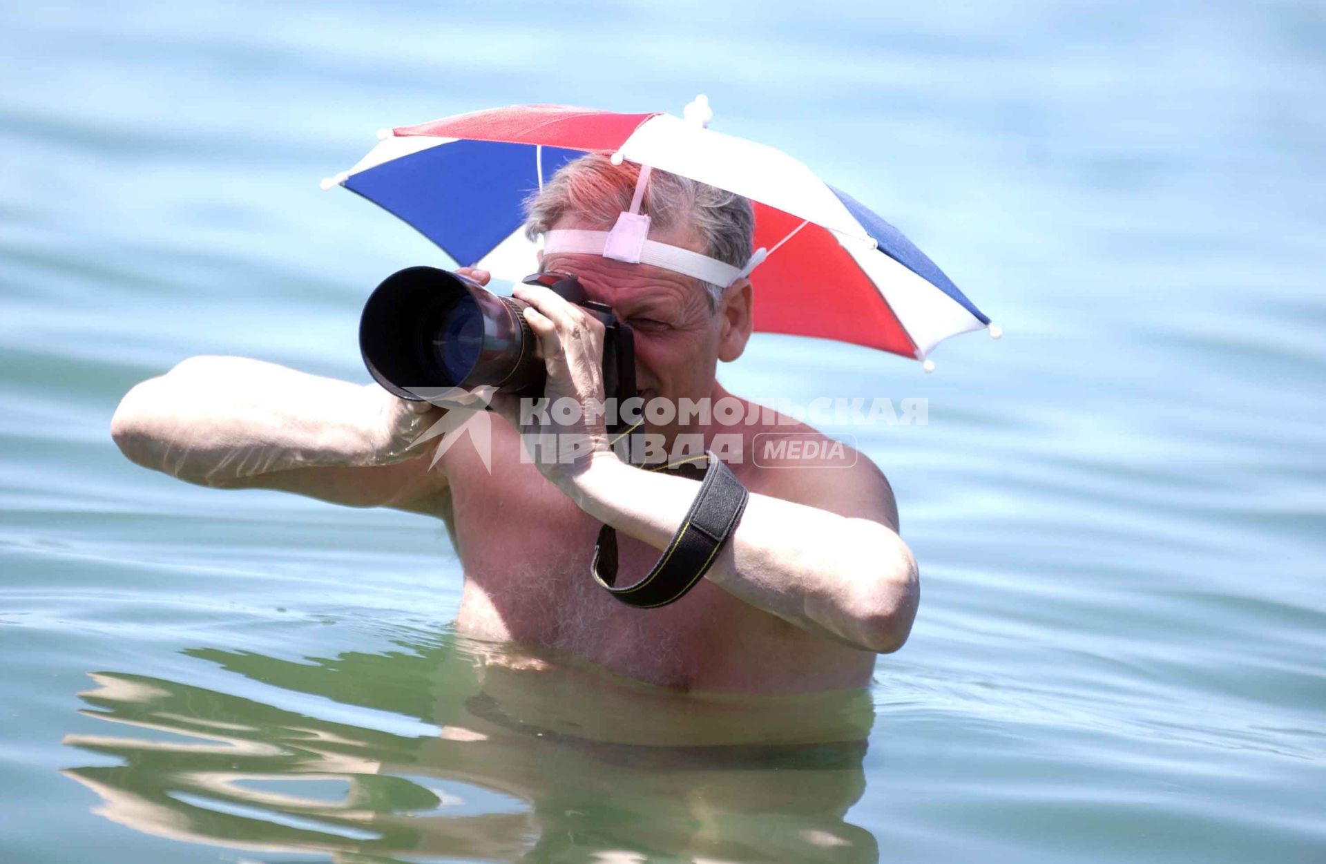 Фотограф снимает стоя в воде с зонтиком на голове.