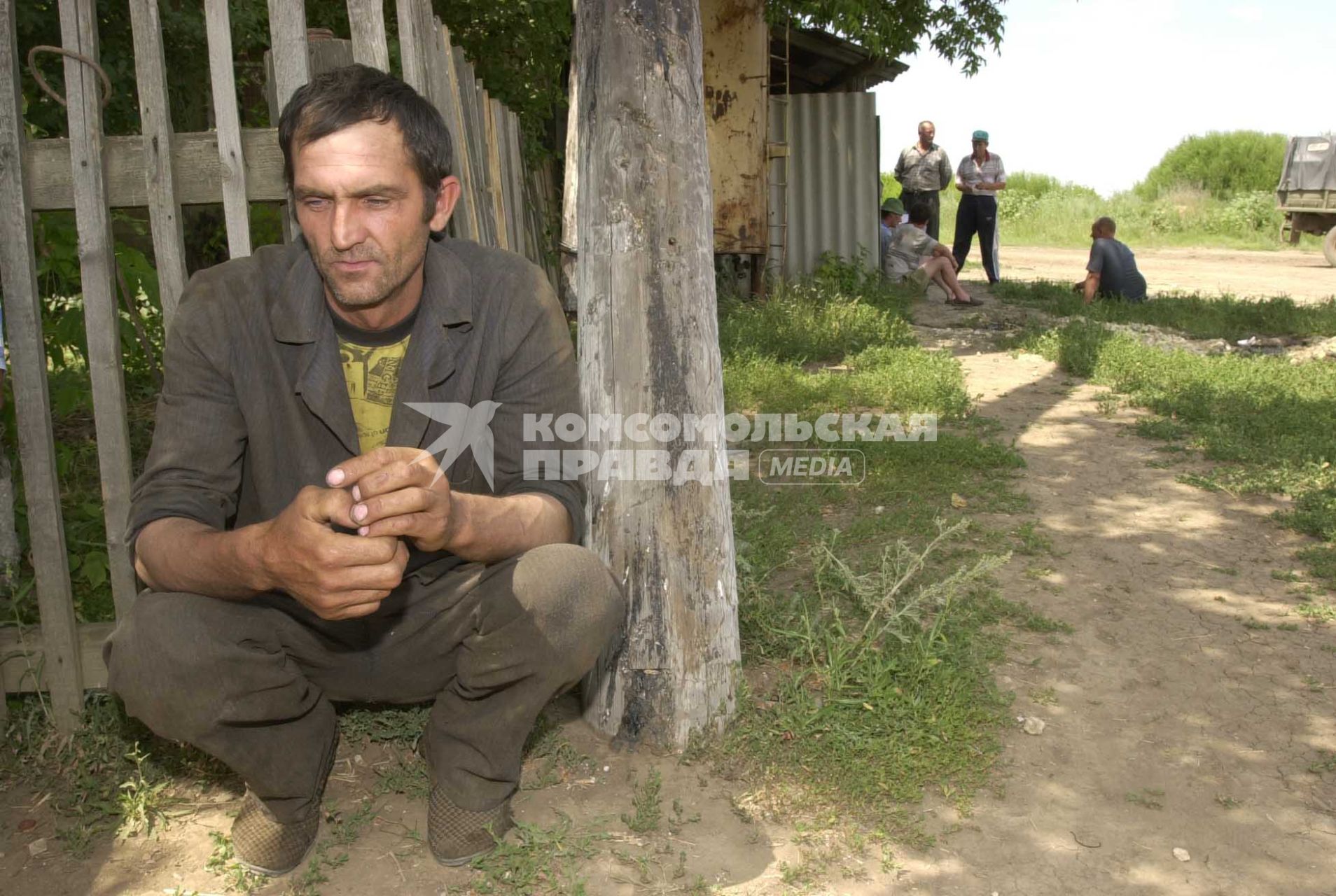 Мужчина в грязной одежде сидит один на корточках, прислонившись к забору, на заднем плане компания мужчин общается между собой.