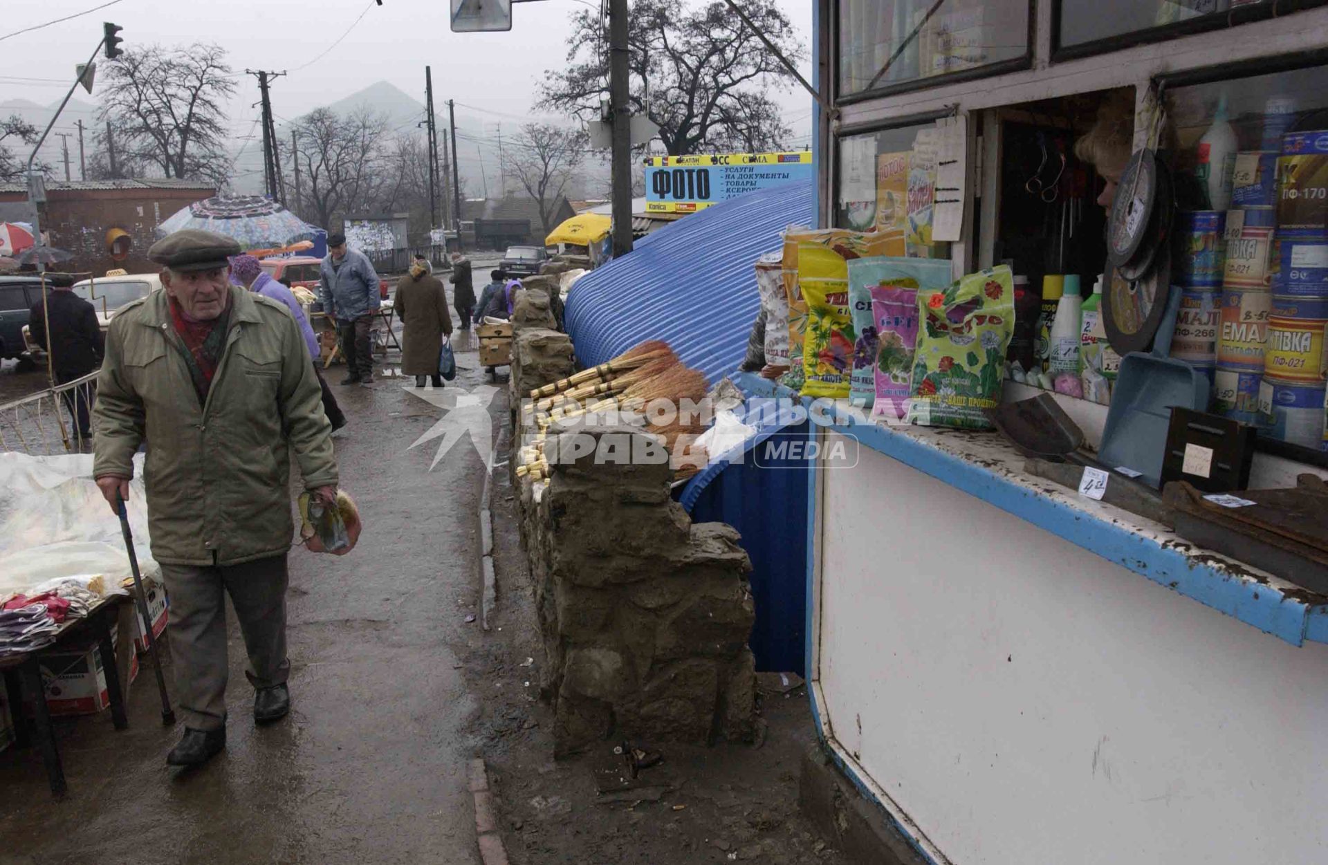 Уличная торговля в провинциальном городе. Пожилой мужчина с костылем проходи мимо палатки хозяйственными товарами.