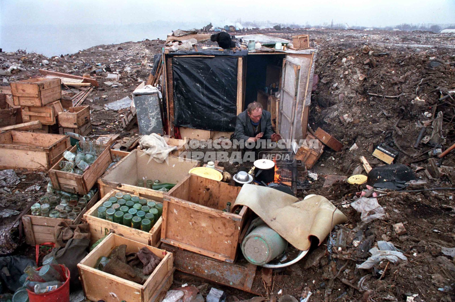 Бродяга греется у костра около своего самодельного дома на свалке.