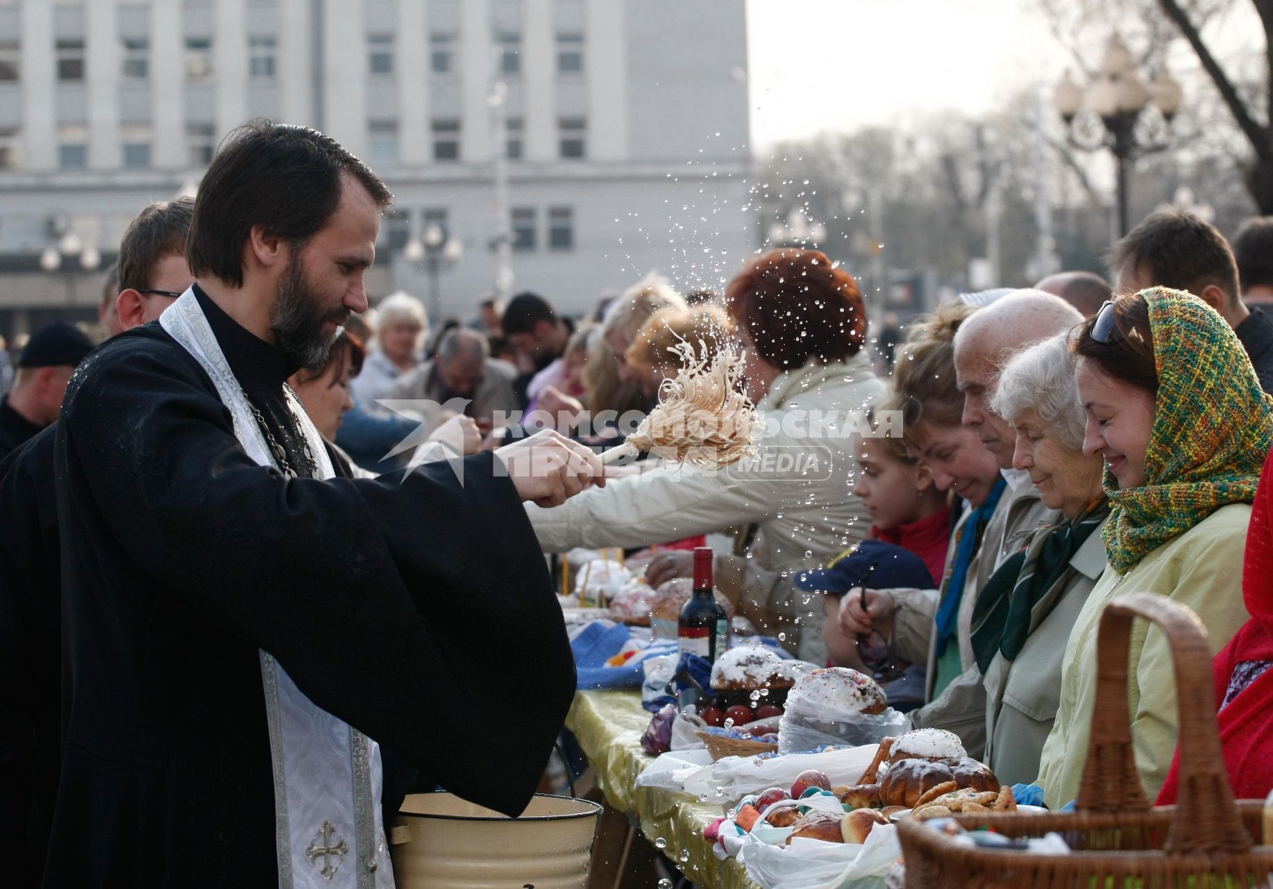 На улице священник освящает куличи, бутылки вина и крашенные яйца обрызгивая кропилом стол с угощениями и прихожан.