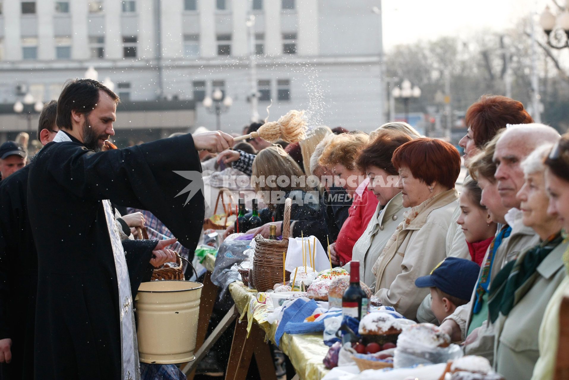 На улице священник освящает куличи, бутылки вина и крашенные яйца обрызгивая кропилом стол с угощениями и прихожан.