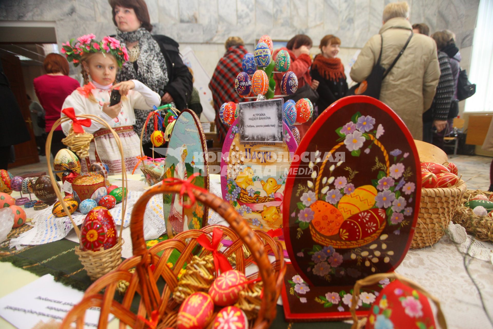 Девочка в национальной одежде фотографирует экспозицию с украшенными пасхальными яйцами.