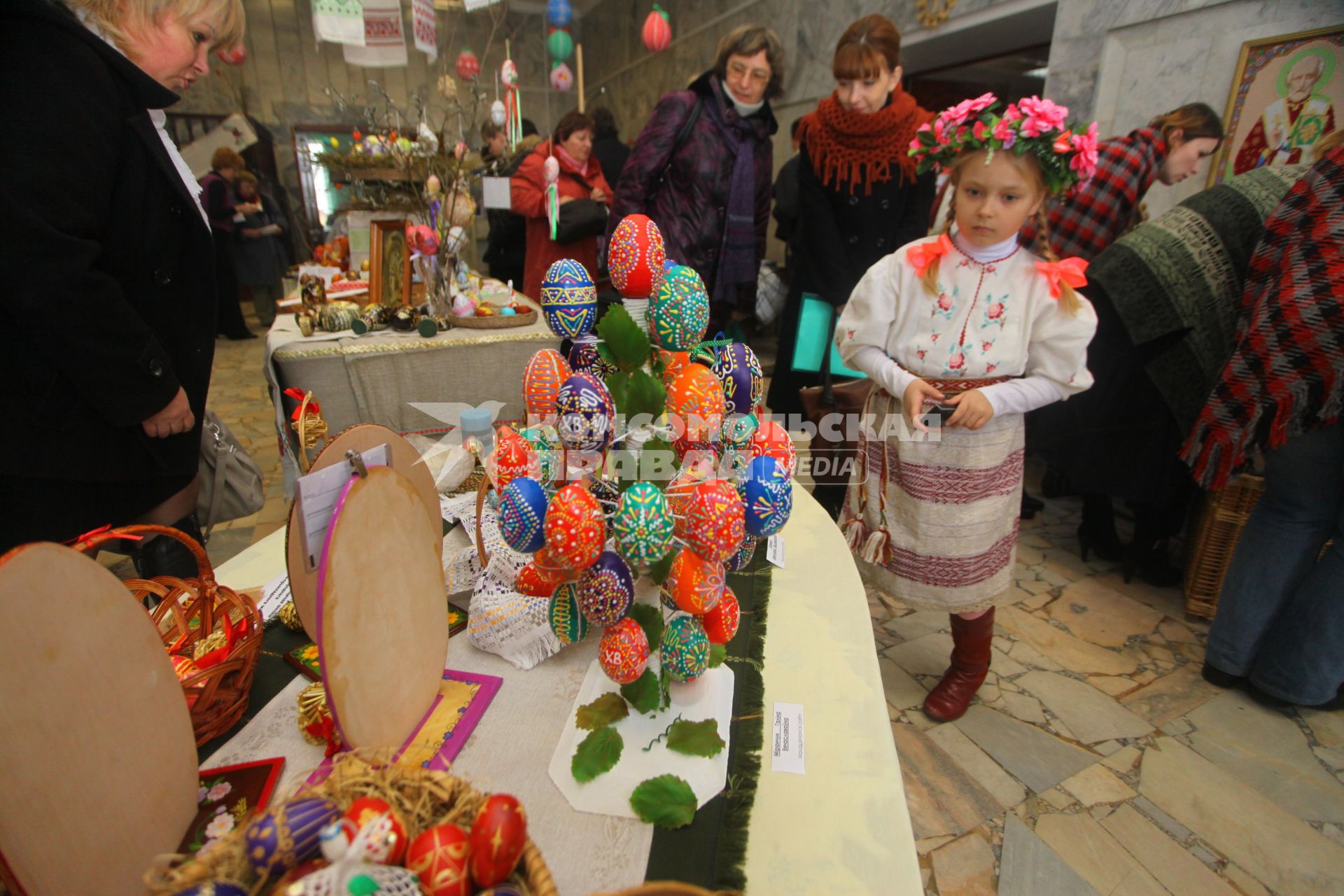 Девочка в национальной одежде стоит рядом со столом на котором стоят пасхальные яйца.