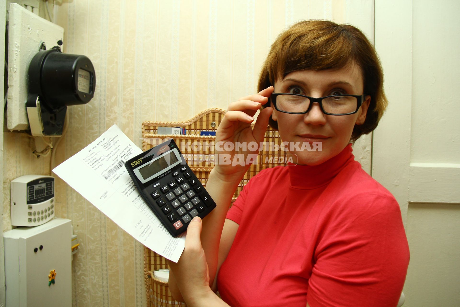 Женщина в очках с калькулятором и квитанцией на оплату коммунальных услуг стоит около счетчика электроэнергии.