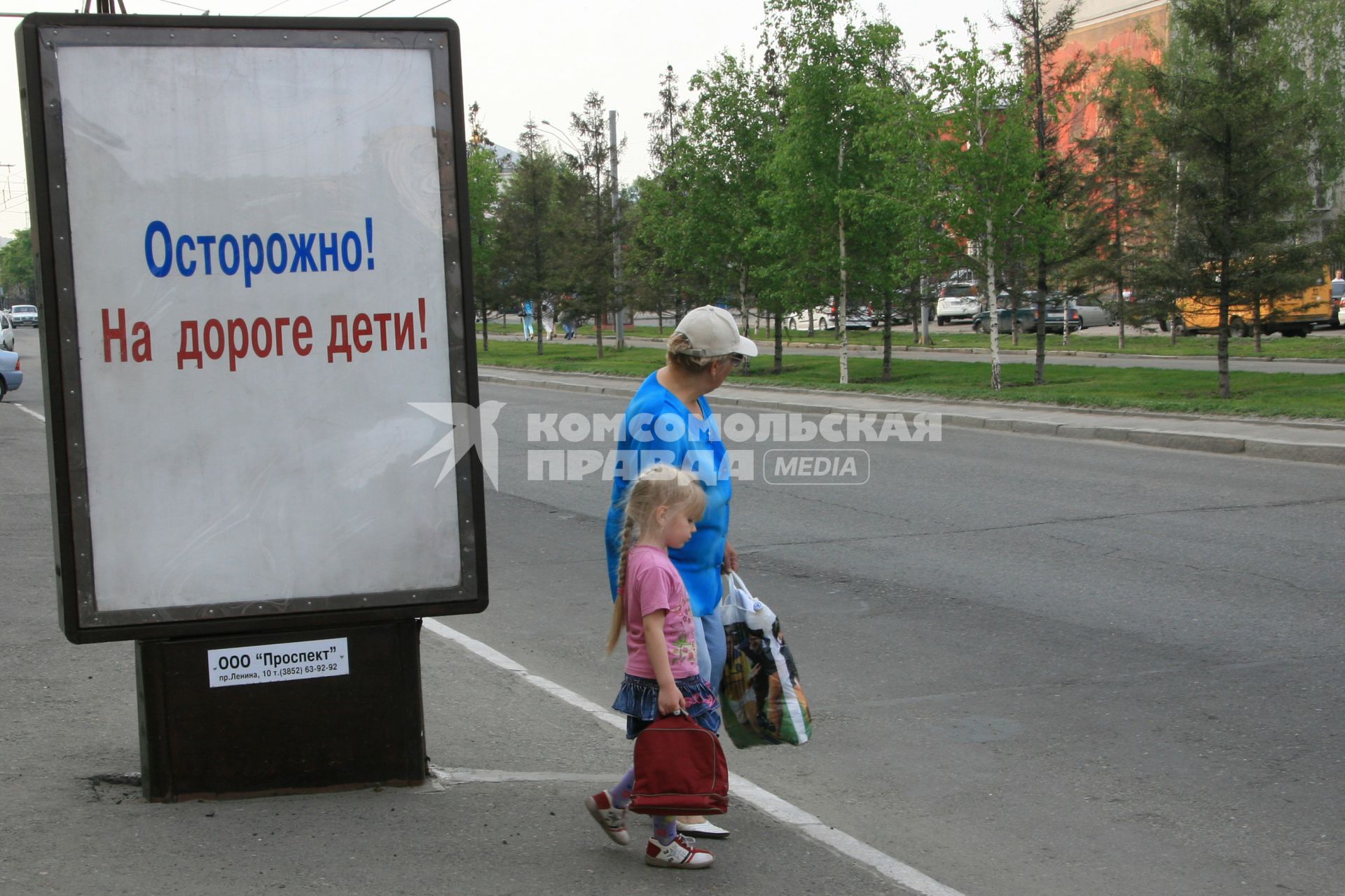 Бабушка с внучкой собираются нарушить правила дорожного движения перейдя автомобильную дорогу в неположенном месте. Рядом стоит рекламный щит с социальной рекламой `Осторожно! На дороге дети!`.