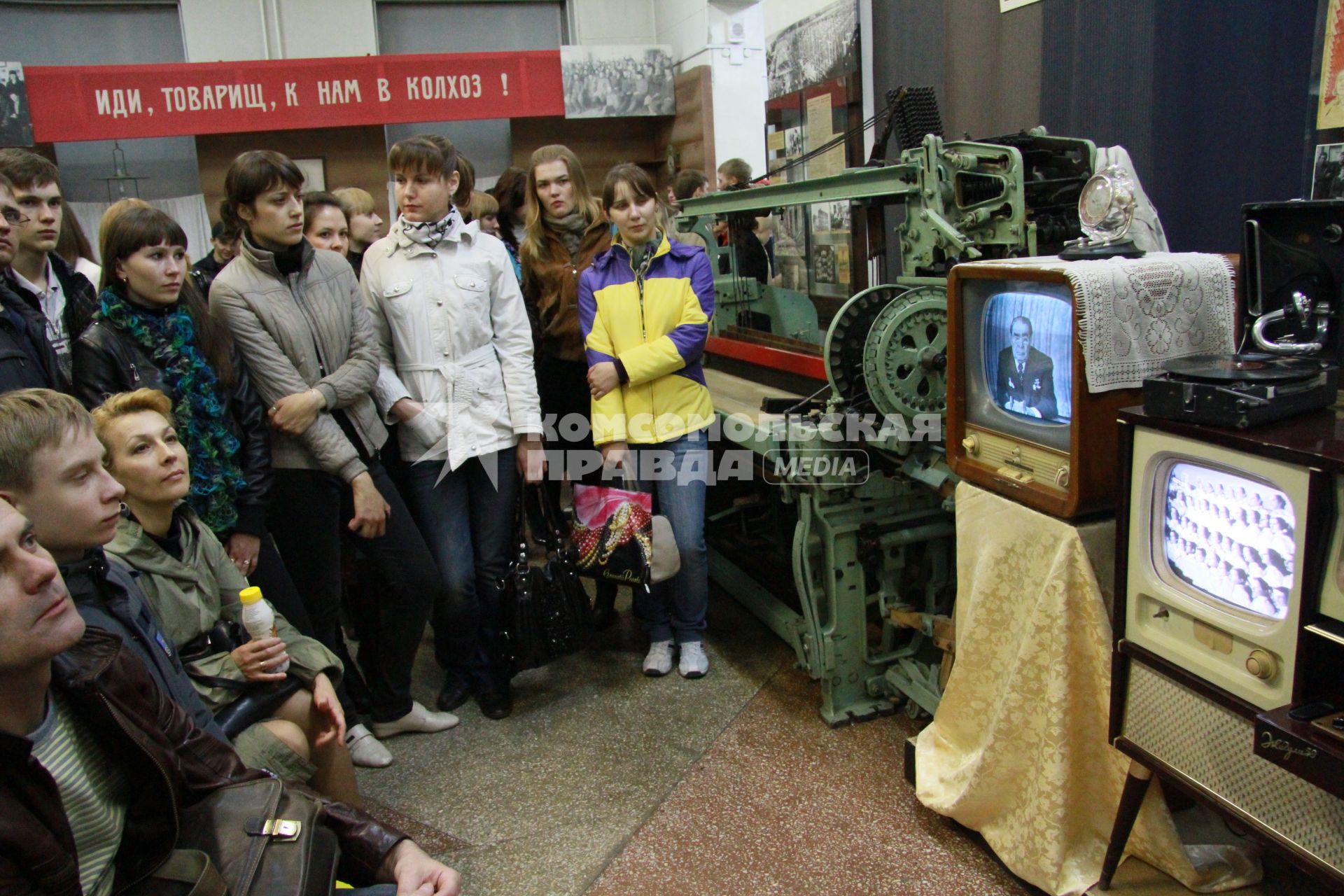 Краеведческий музей. Посетители смотрят телевизионные программы времен СССР по черно-белым телевизорам. Сзади плакат с надписью: `Иди, товарищ, к нам в колхоз!`.