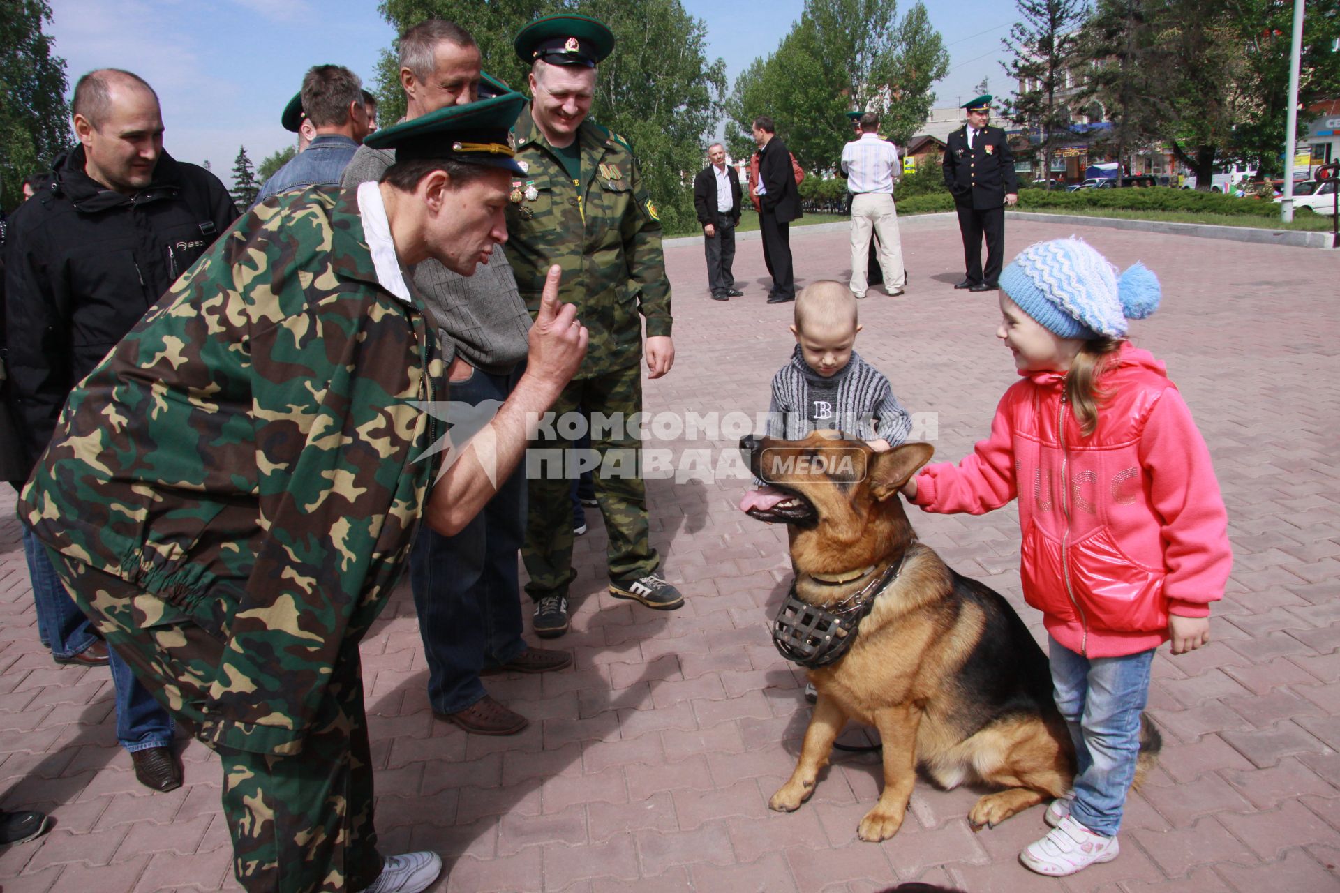 Мужчина в форме пограничника что-то рассказывает собаке с намордником, которую за ухи трепят двое смеющихся детей.