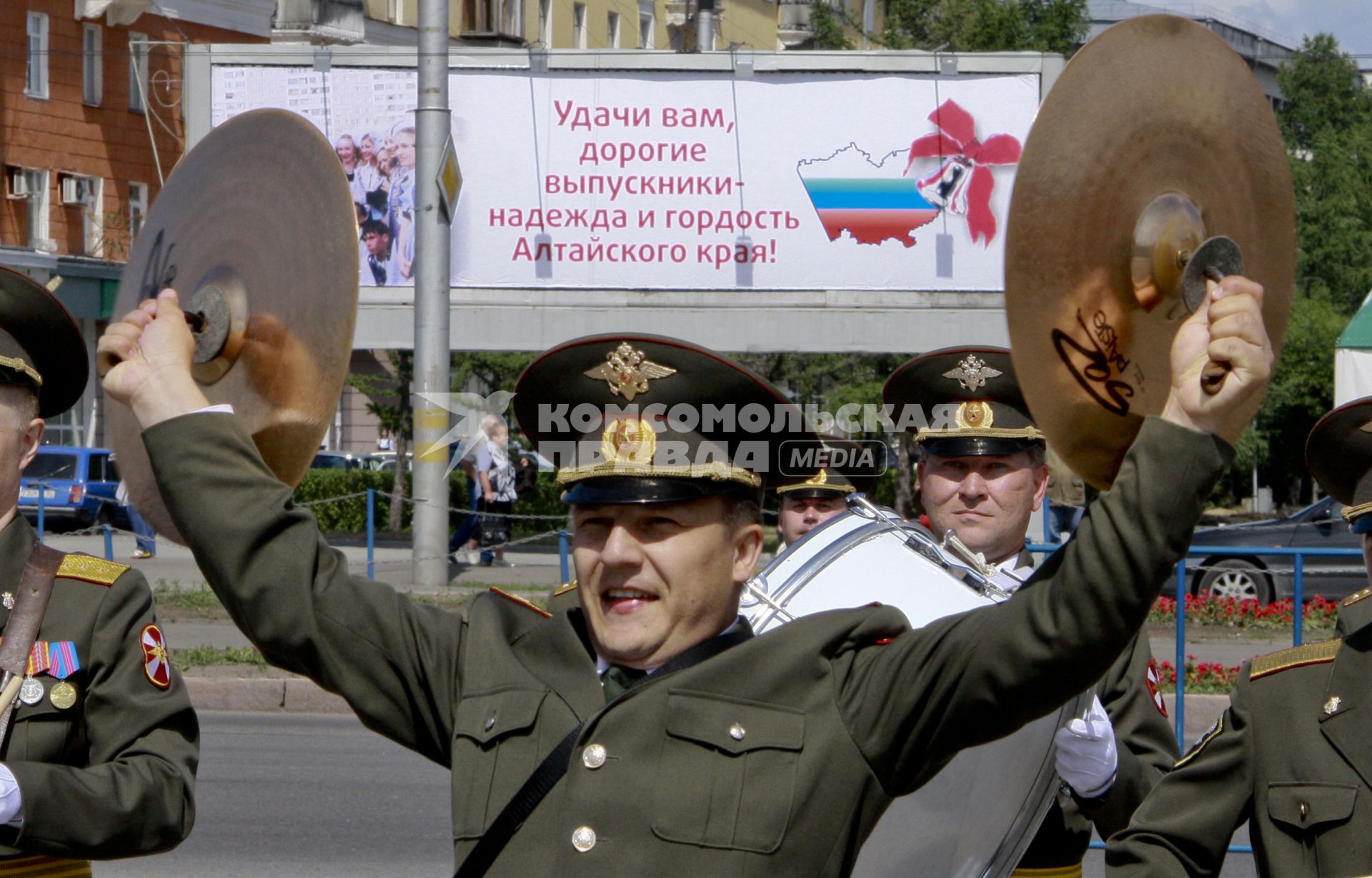 Военный оркестр играет музыку на улице города, сзади плакат с текстом: `Удачи вам, дорогие выпускники - надежда и гордость Алтайского края!`