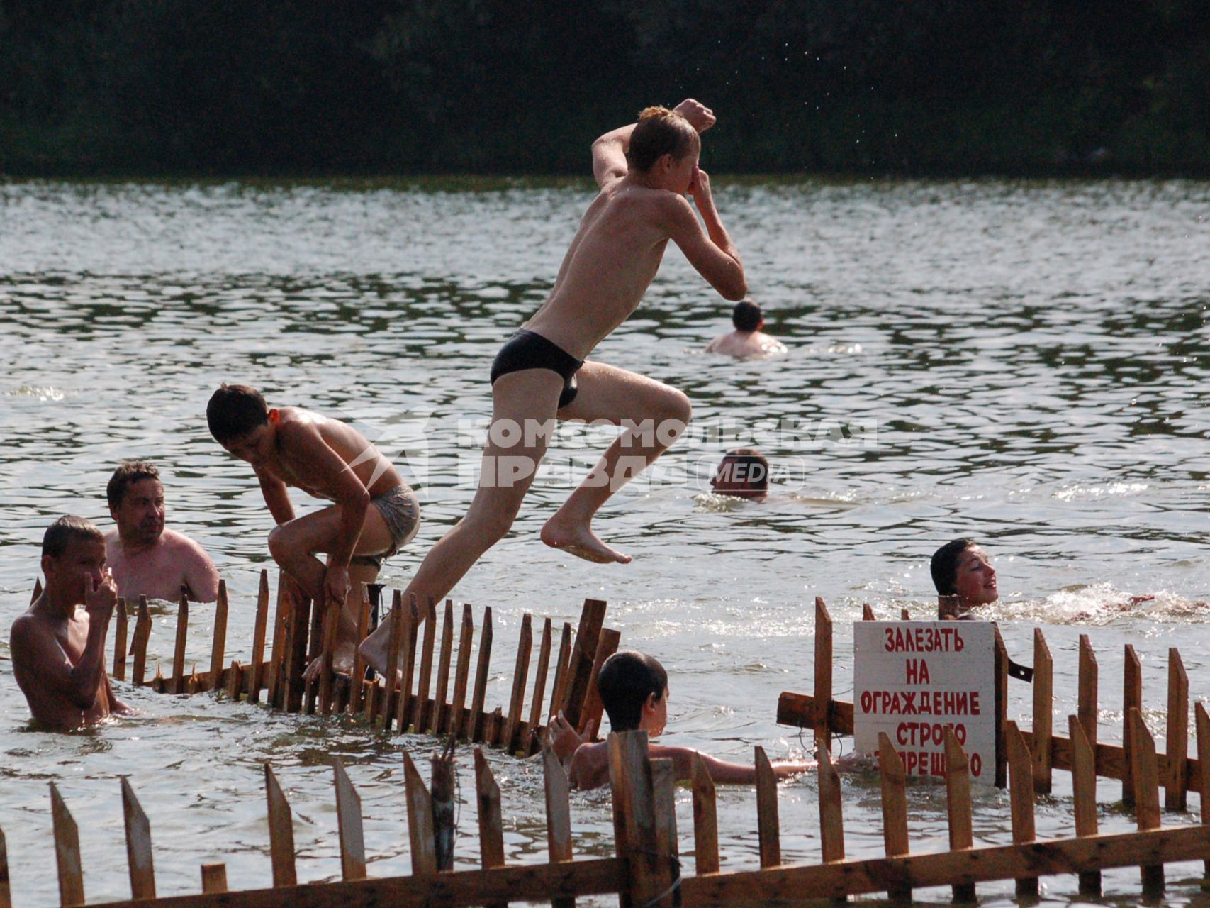 Мальчик прыгает в воду с ограждения на котором прикреплена надпись: `Залезать на ограждение строго запрещено`.