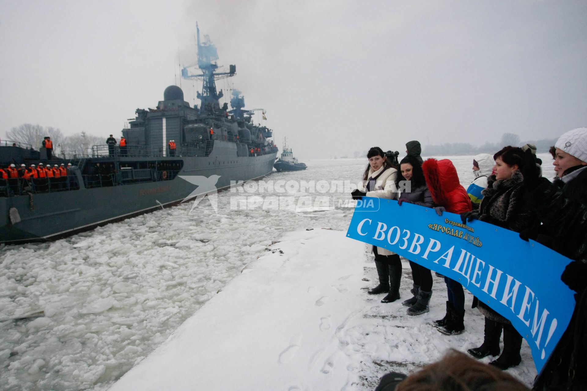 Сторожевик Балтфлота `Ярослав Мудрый` вернулся в порт 14 февраля. Люди стоят на пристане с плакатом: `С возвращением!`.