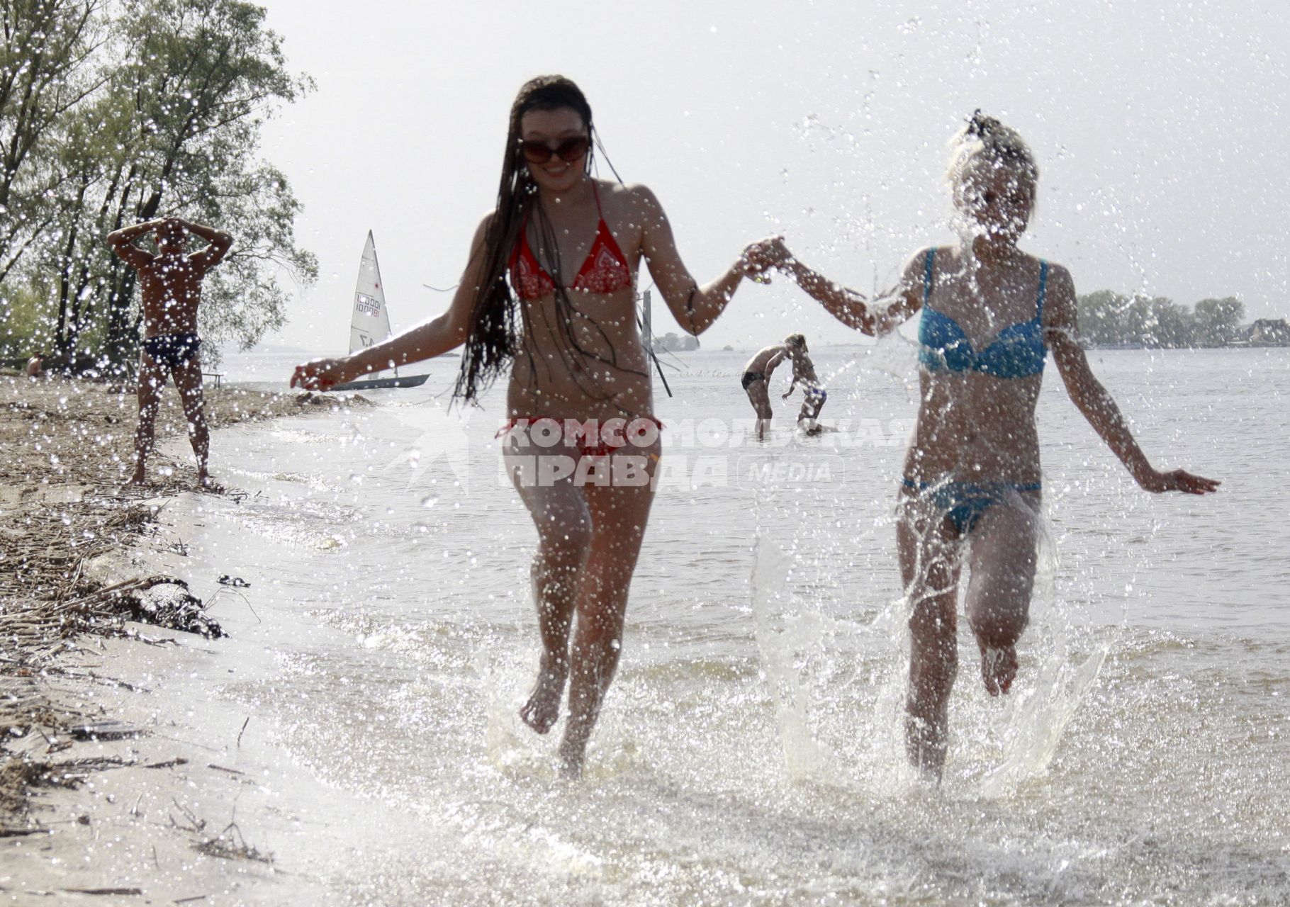 На пляже две девушки бегут с брызгами по воде.