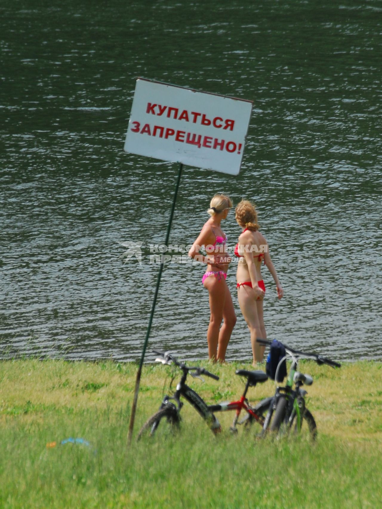 Две девушки в купальниках стоят на берегу водоема, рядом табличка `Купаться запрещено`.