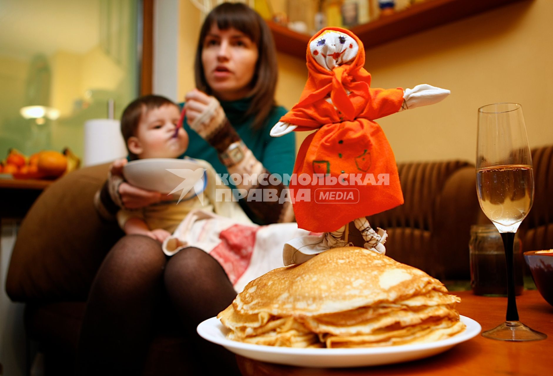 Девушка кормит ребенка на фоне тарелки с блинами и чучела масленицы.