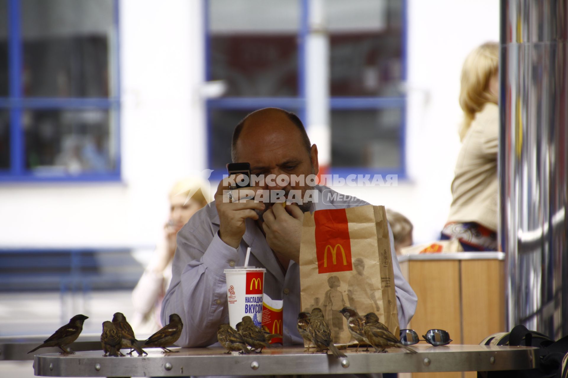 Мужчина питается в уличном кафе `Макдональдса`. На стол к нему прилетели стая воробьев.