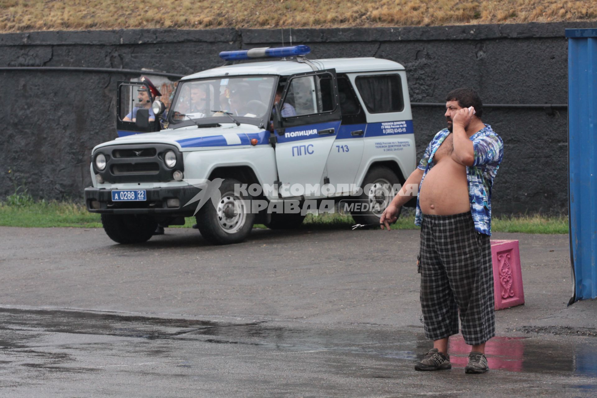 Мужчина в расстегнутоий рубашке с большим голым животом говорит по телефону на фоне машины полиции.