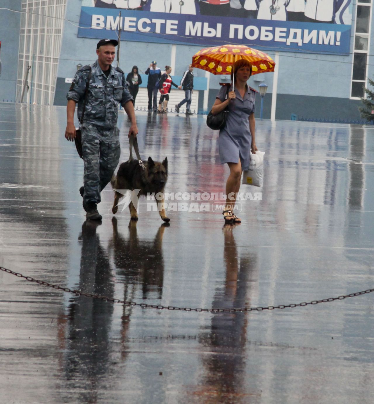 Кинолог с собакой и женщина в полицейской форме под дождем на фоне надписи: `Вместе мы победим`.