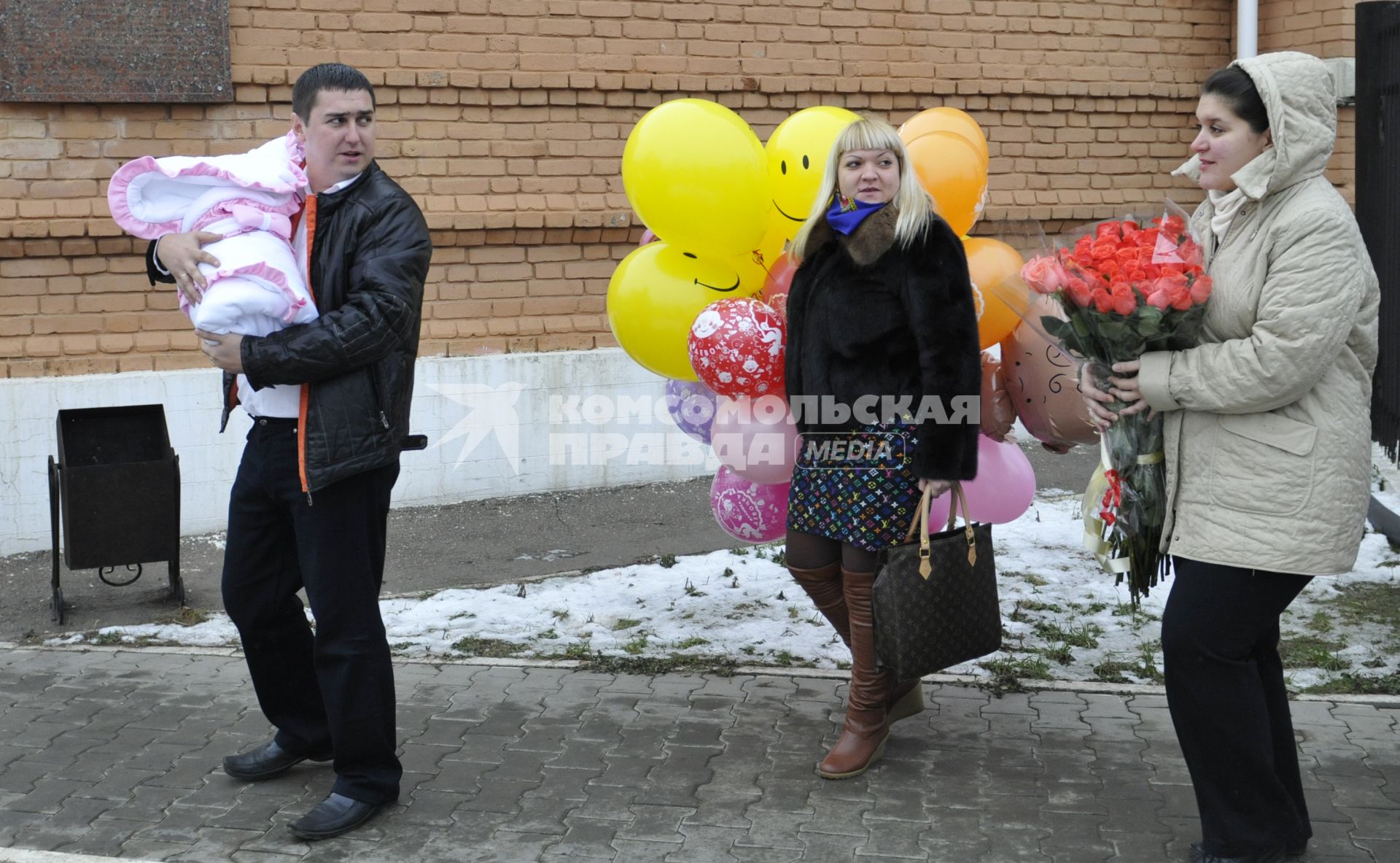 Высписка новорожденного с мамой из роддома. На снимке: Отец несет ребенка в конверте сзади идет женщина с воздушными шарами и мать с букетом цветов из роз.