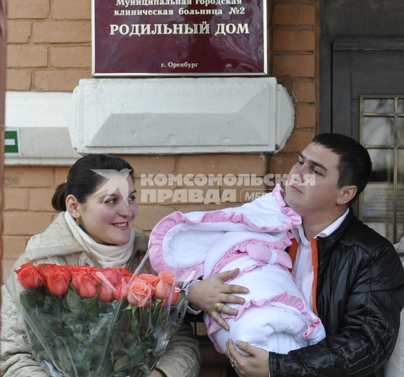 Высписка новорожденного с мамой из роддома. На снимке: Счастливые родители перед роддомом. Отец держит ребенка завернутого в конверт, мать держит букет цветов из роз.