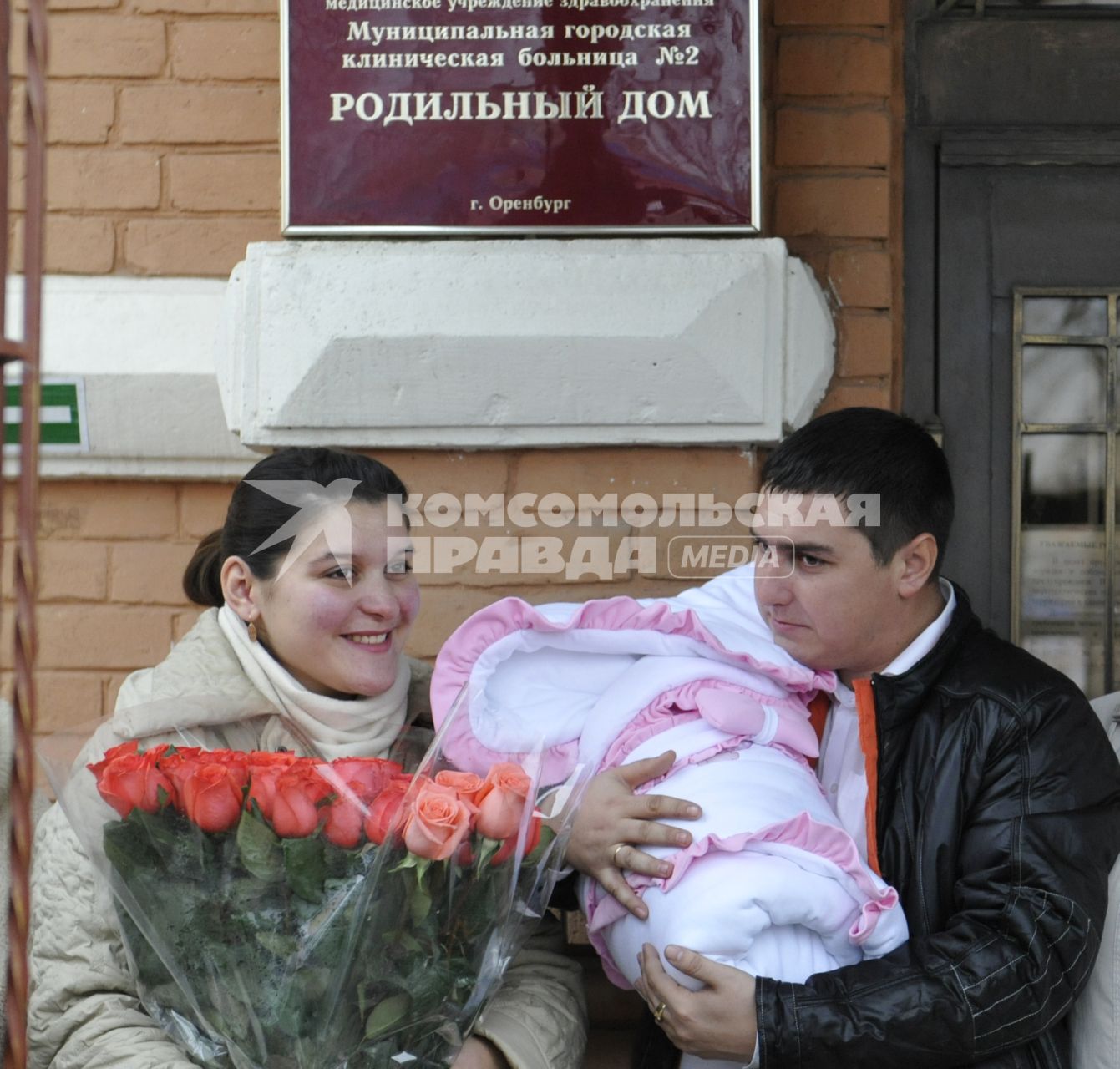 Высписка новорожденного с мамой из роддома. На снимке: Счастливые родители перед роддомом. Отец держит ребенка завернутого в конверт, мать держит букет цветов из роз.