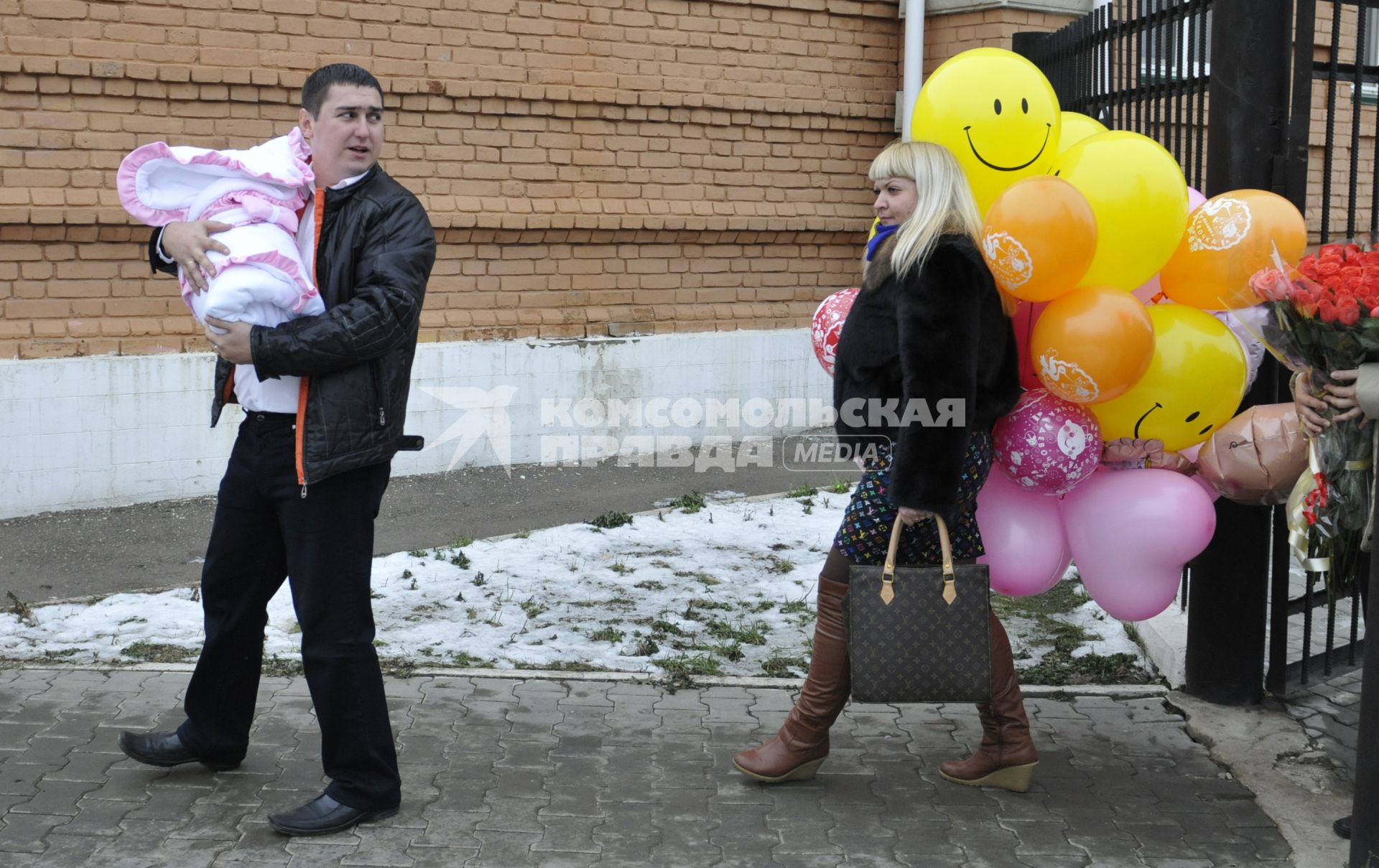 Высписка новорожденного с мамой из роддома. На снимке: Отец несет ребенка в конверте сзади идет женщина с воздушными шарами.