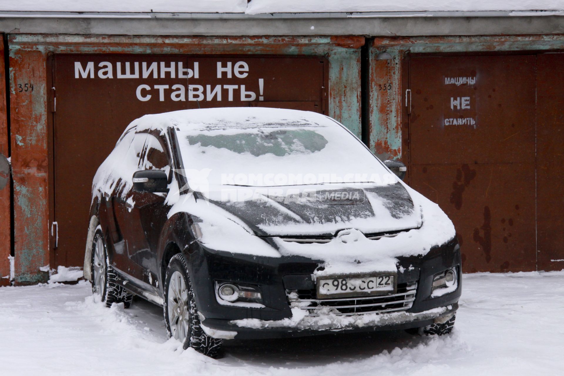 Перед гаражом с надписью `Машины не ставить` стоит заснеженный автомобиль.