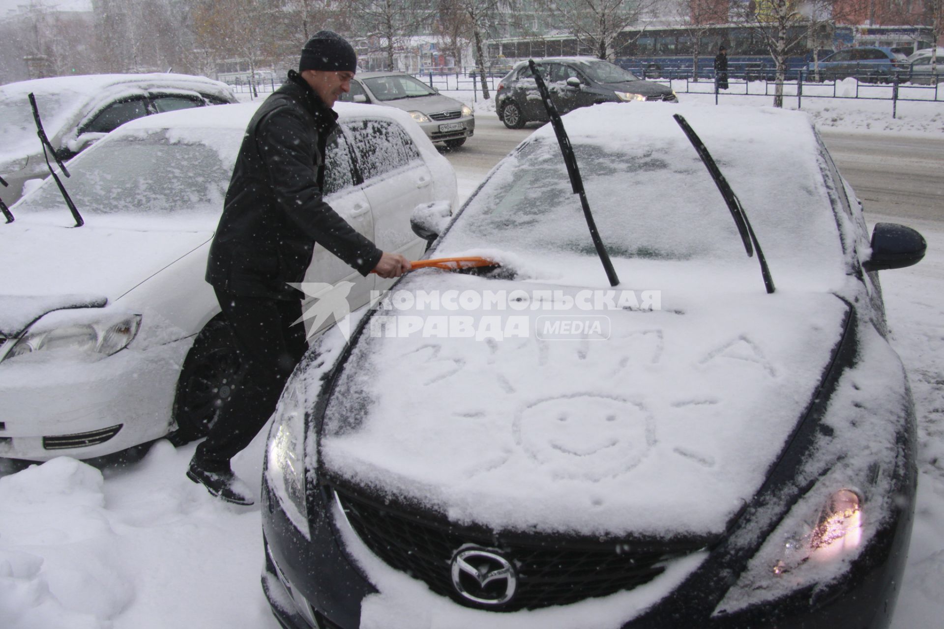 Водитель чистит автомобиль от снега. На капоте надпись `зима`.