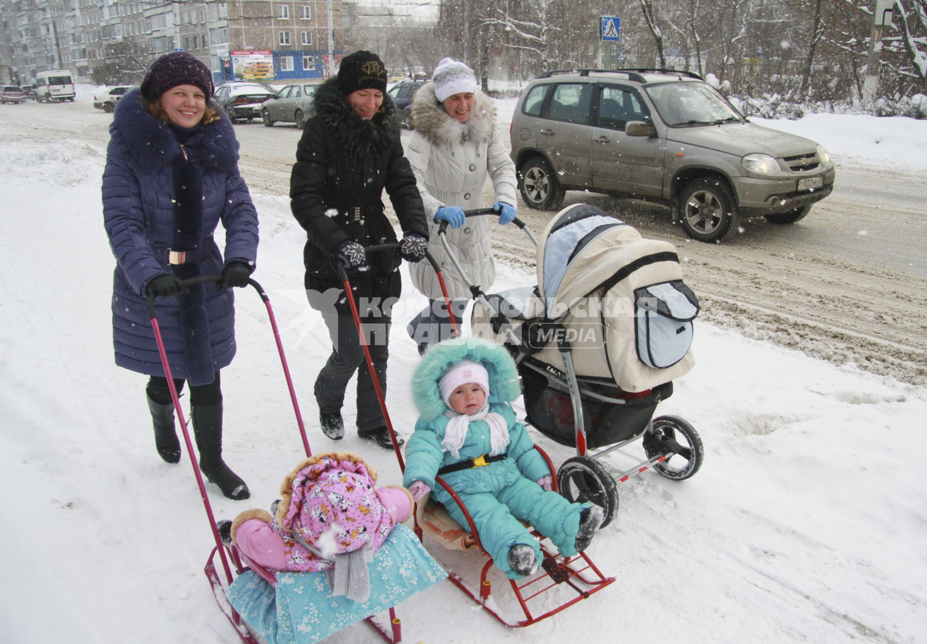 Три женщины гуляют с детьми по городу. Двое детей едут на санках, одного везут в детской коляске.