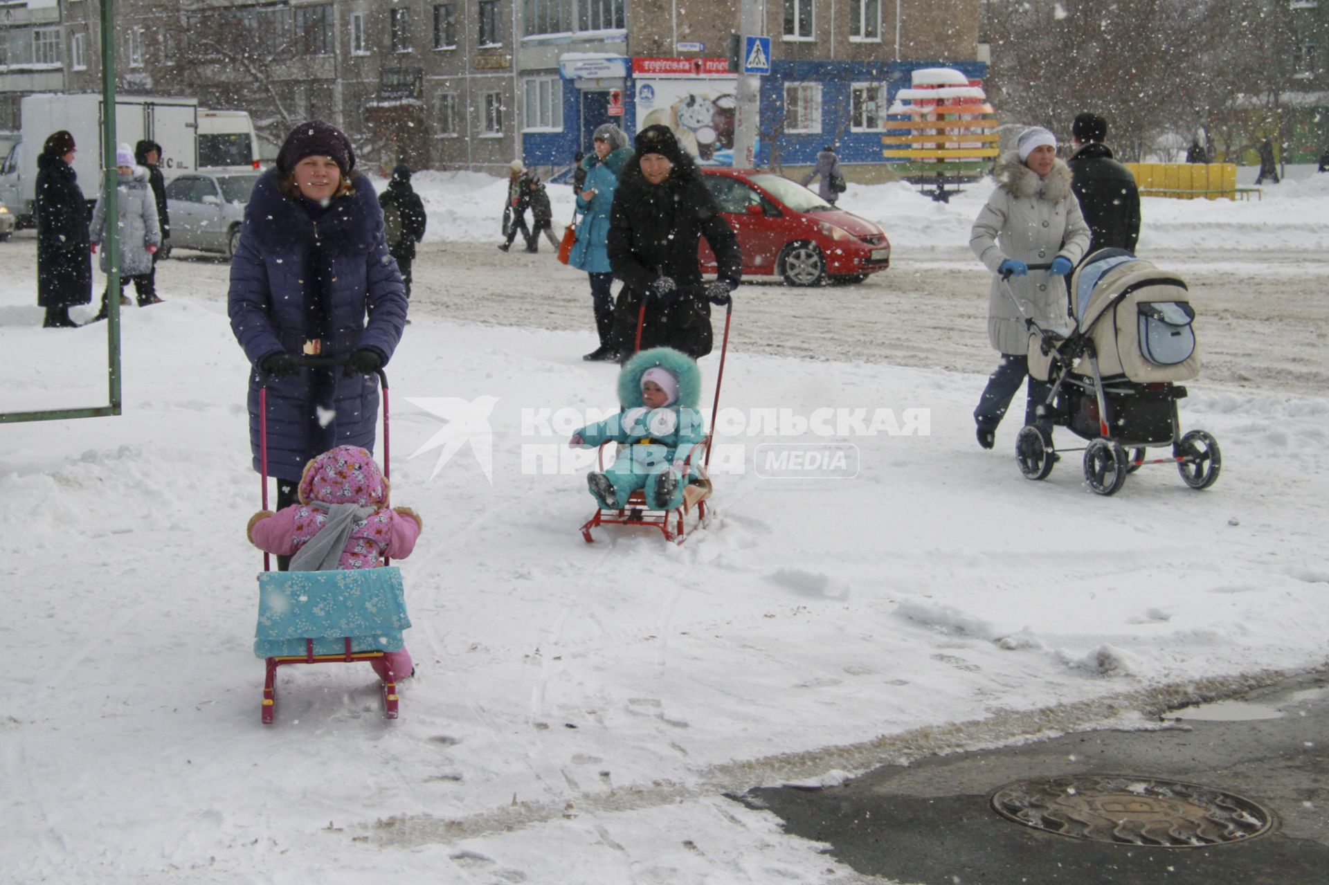 Три женщины гуляют с детьми по городу. Двое детей едут на санках, одного везут в детской коляске.
