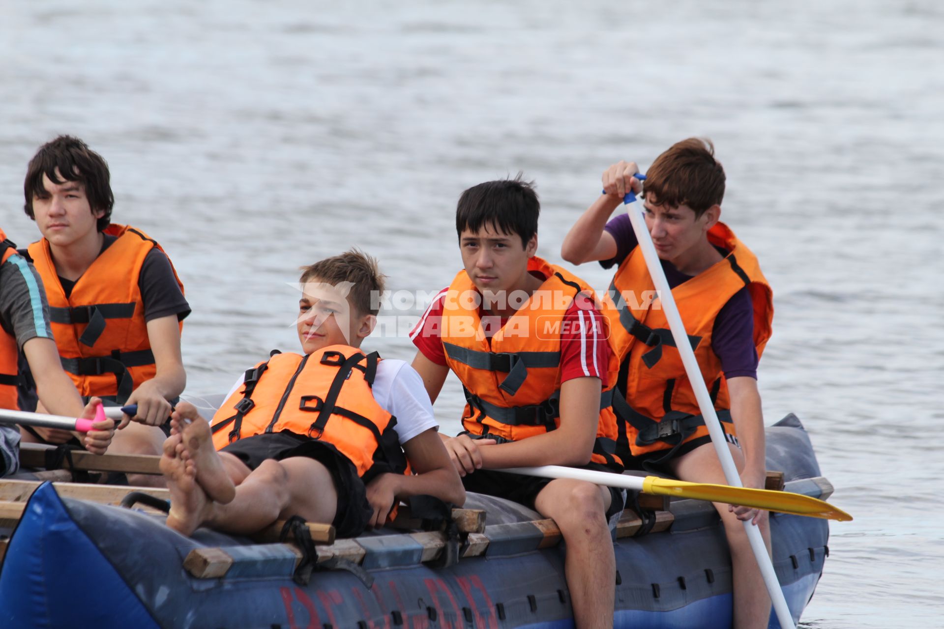 Группа молодых ребят в спасательных жилетах катается на надувном катамаране с веслами.