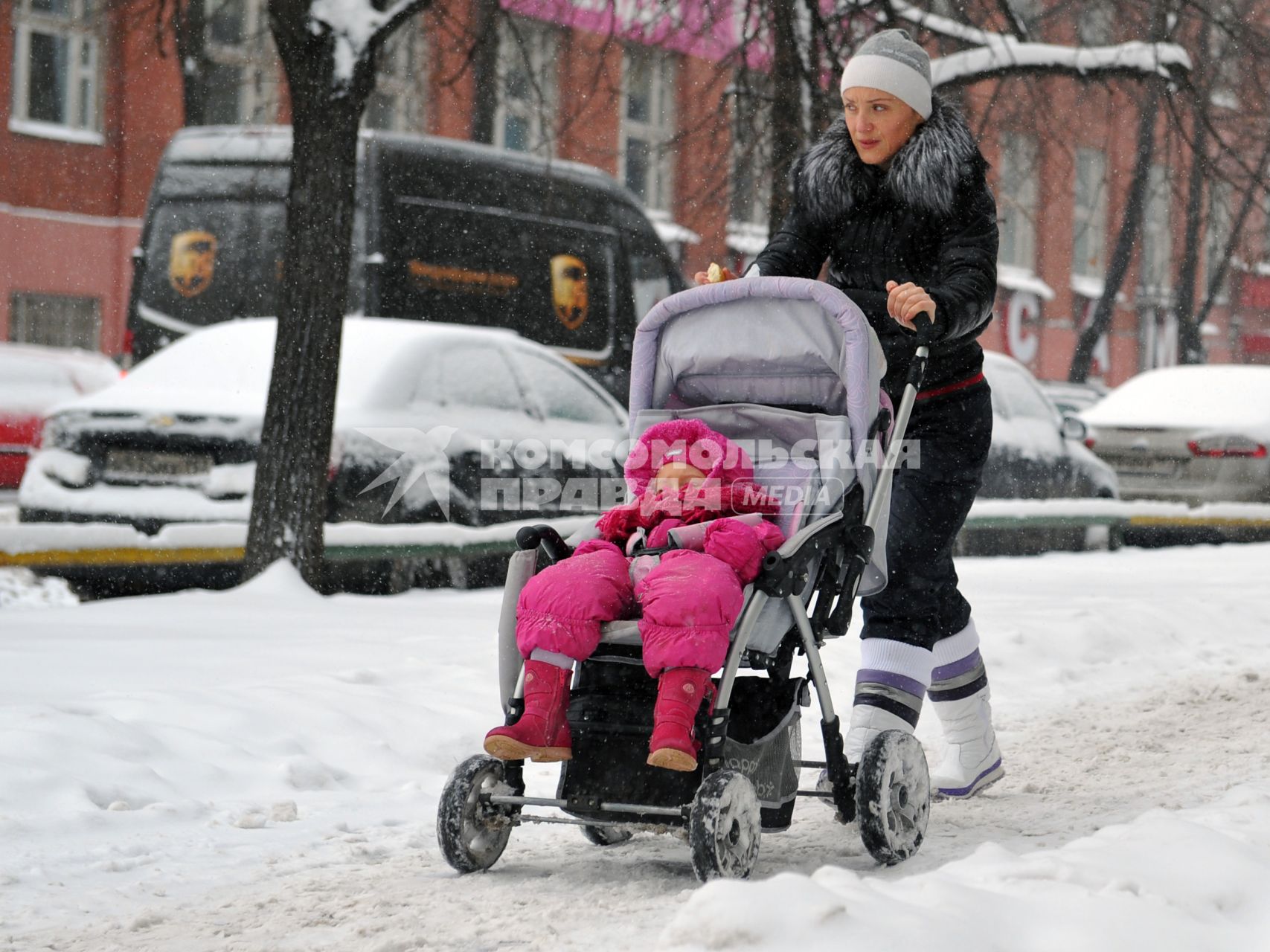 Женщина с ребенком в детской коляске идет по заснеженной улице.