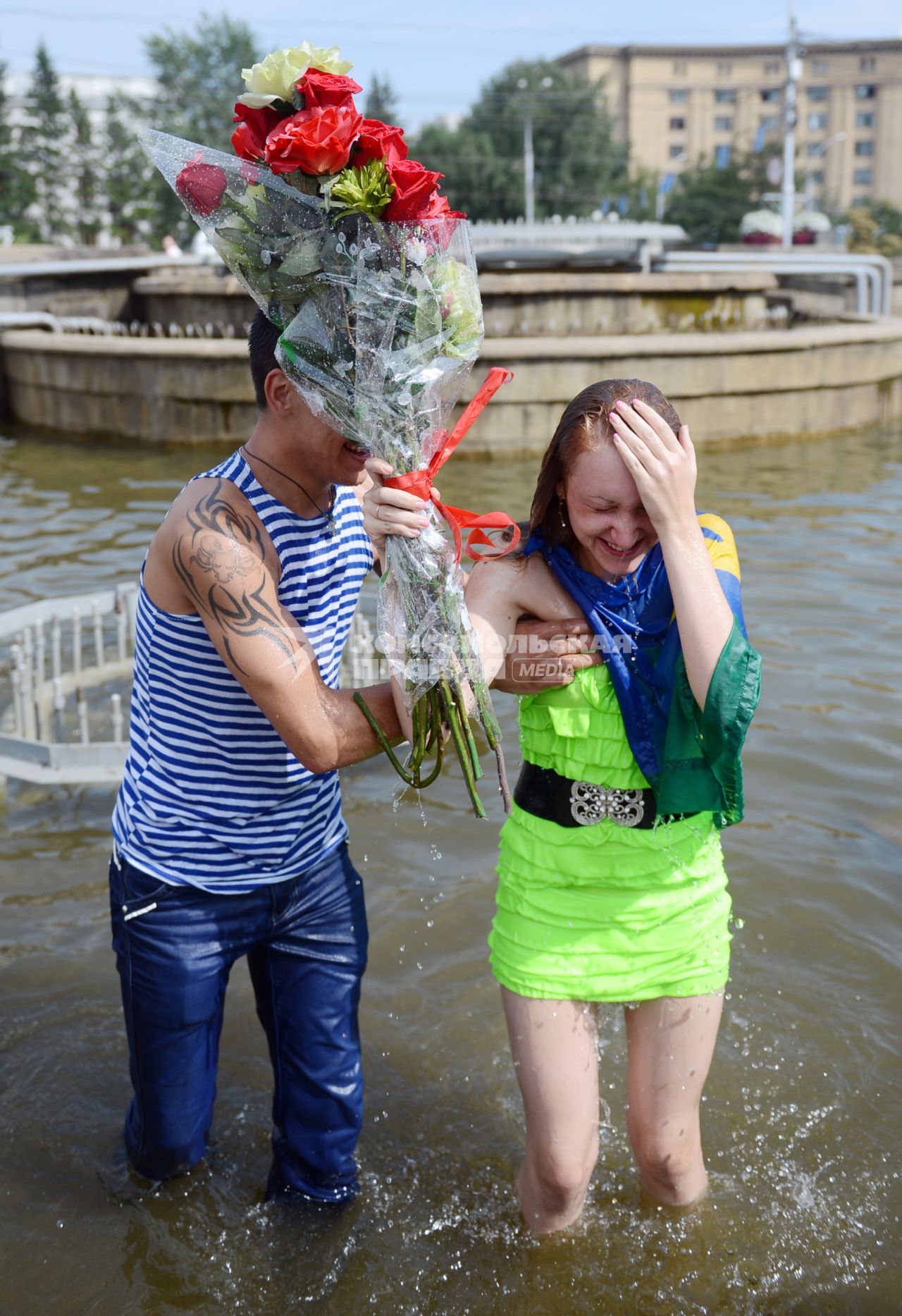 Мужчина в тельняшке десантника помогает выбраться из фонтана девушке с букетом цветов.